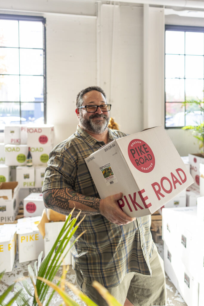 Smiling bearded man in dark rimmed glasses with a green plaid shirt carrying a case of Pike Road wine